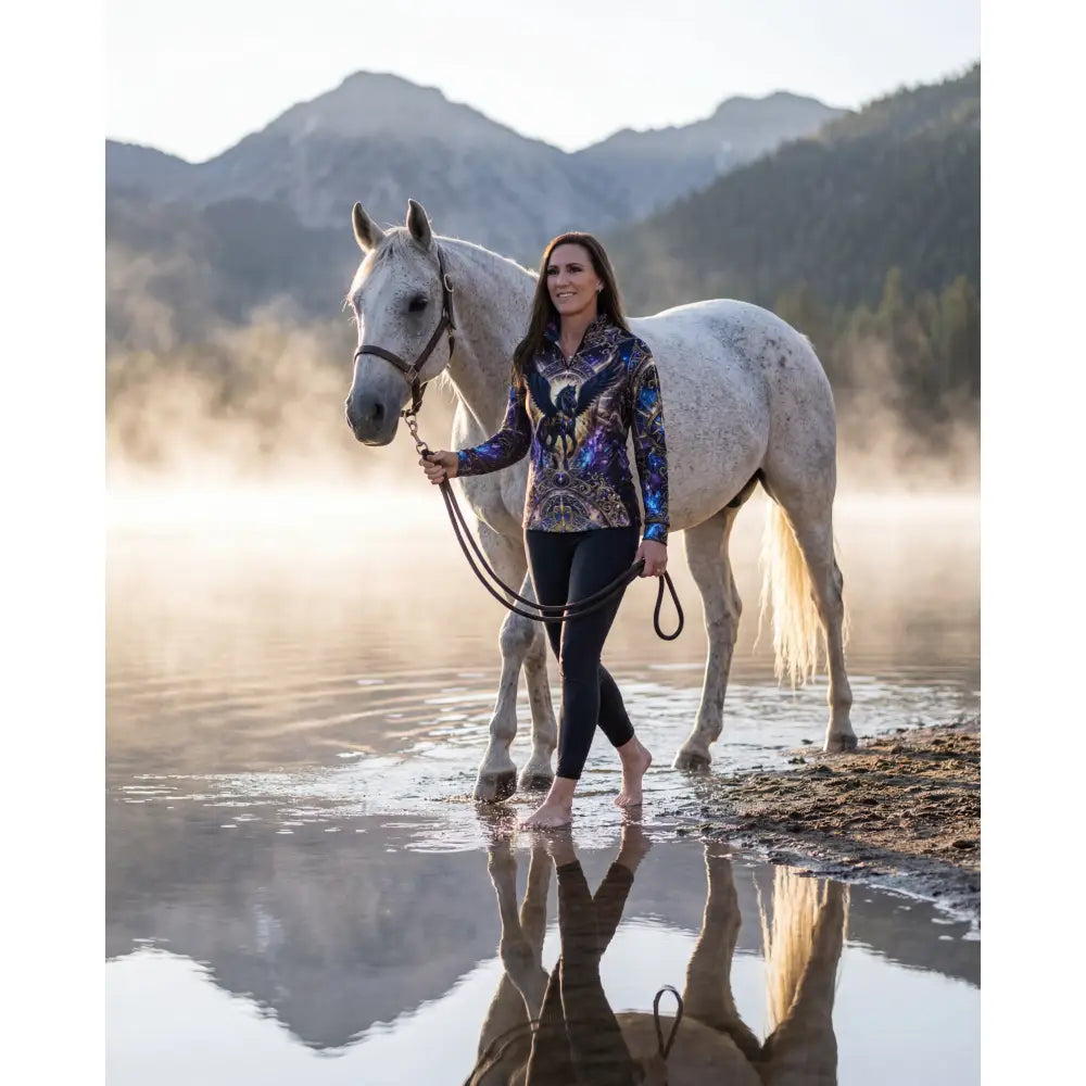 A woman wearing an astral ascension galaxy-patterned performance equestrian sun shirt stands beside a dapple-gray horse