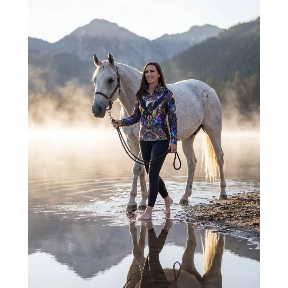 A woman in a vibrant galaxy-patterned performance equestrian sun shirt with upf50+ stands barefoot by a dapple gray horse