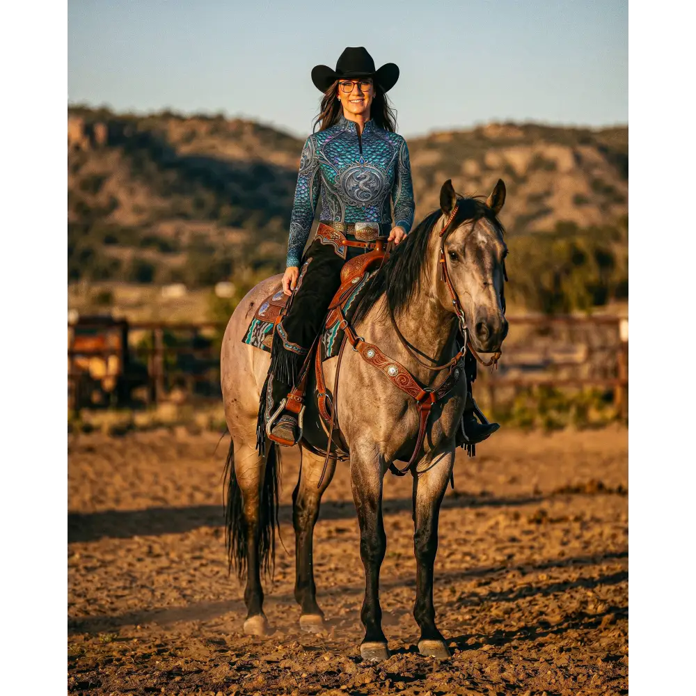 A woman wearing a black cowboy hat rides a gray horse while wearing a blue patterned Caelestia Draconis performance equestrian sun shirt UPF50+