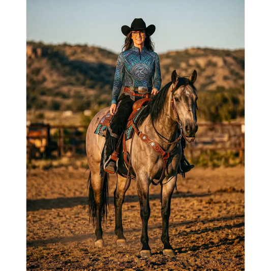 A woman wearing a black cowboy hat rides a gray horse while wearing a blue patterned Caelestia Draconis performance equestrian sun shirt UPF50+