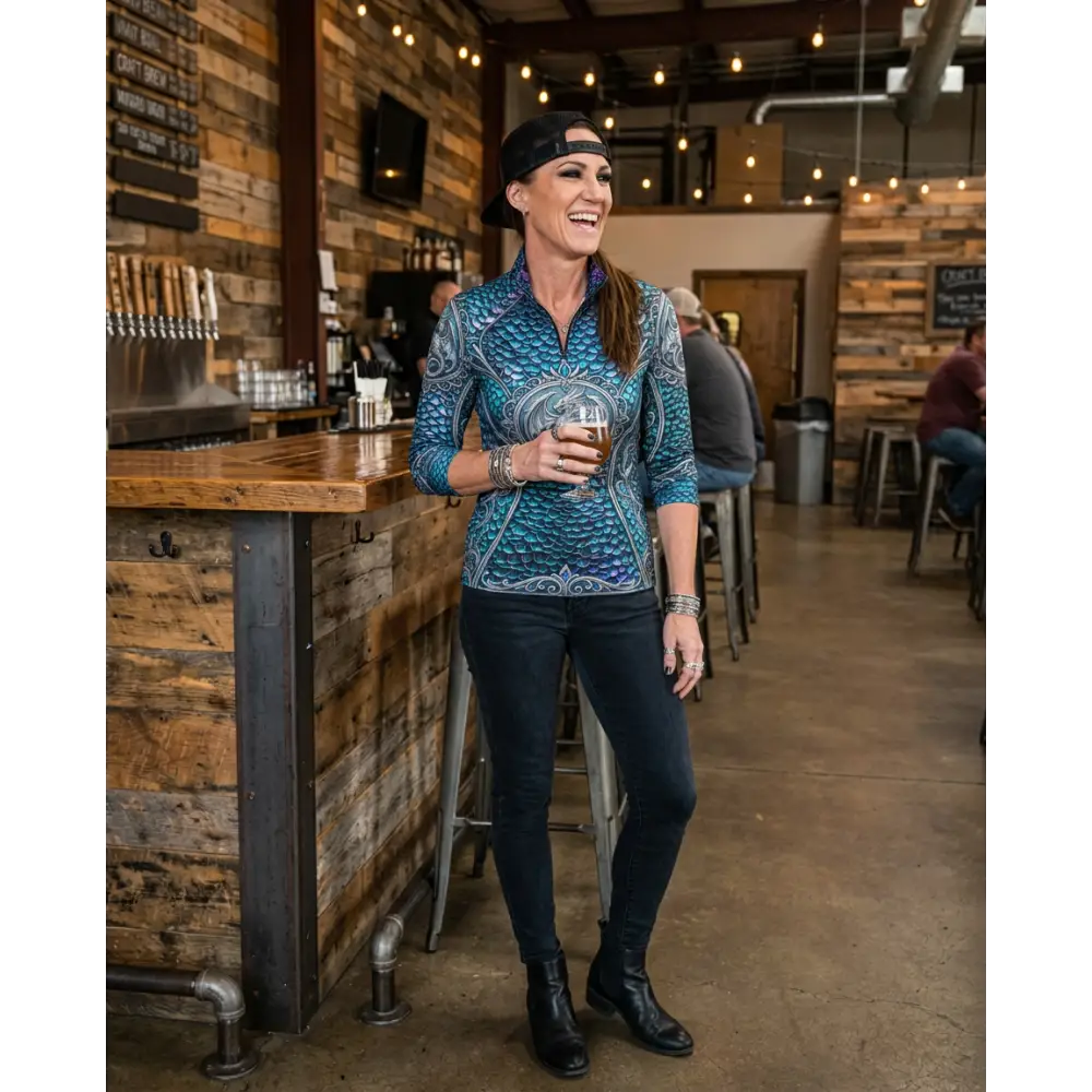 A woman wears a teal and blue patterned Caelestia Draconis performance equestrian sun shirt with intricate scale designs and UPF50+ protection