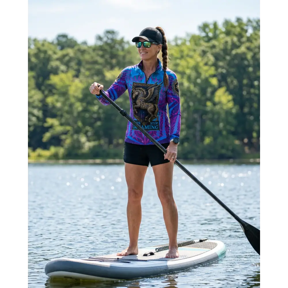 A woman stands on a white and gray inflatable paddleboard wearing a never stop dreaming performance equestrian sun shirt upf50+