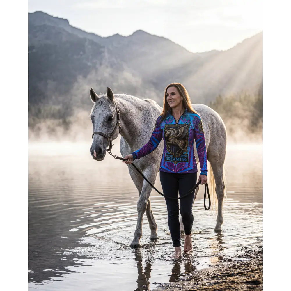 A woman walks barefoot beside a dappled gray horse wearing a vibrant long-sleeved Never Stop Dreaming sun shirt upf50+
