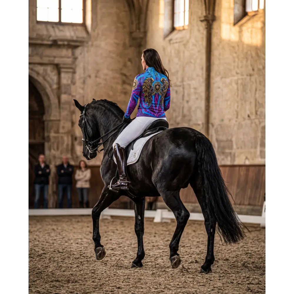 A rider wearing a purple and blue never stop dreaming performance equestrian sun shirt upf50 plus with gold wing designs on a black horse