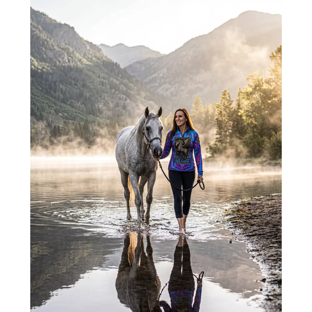 A woman walks barefoot beside a dappled gray horse wearing a vibrant patterned never stop dreaming performance equestrian sun shirt upf50+