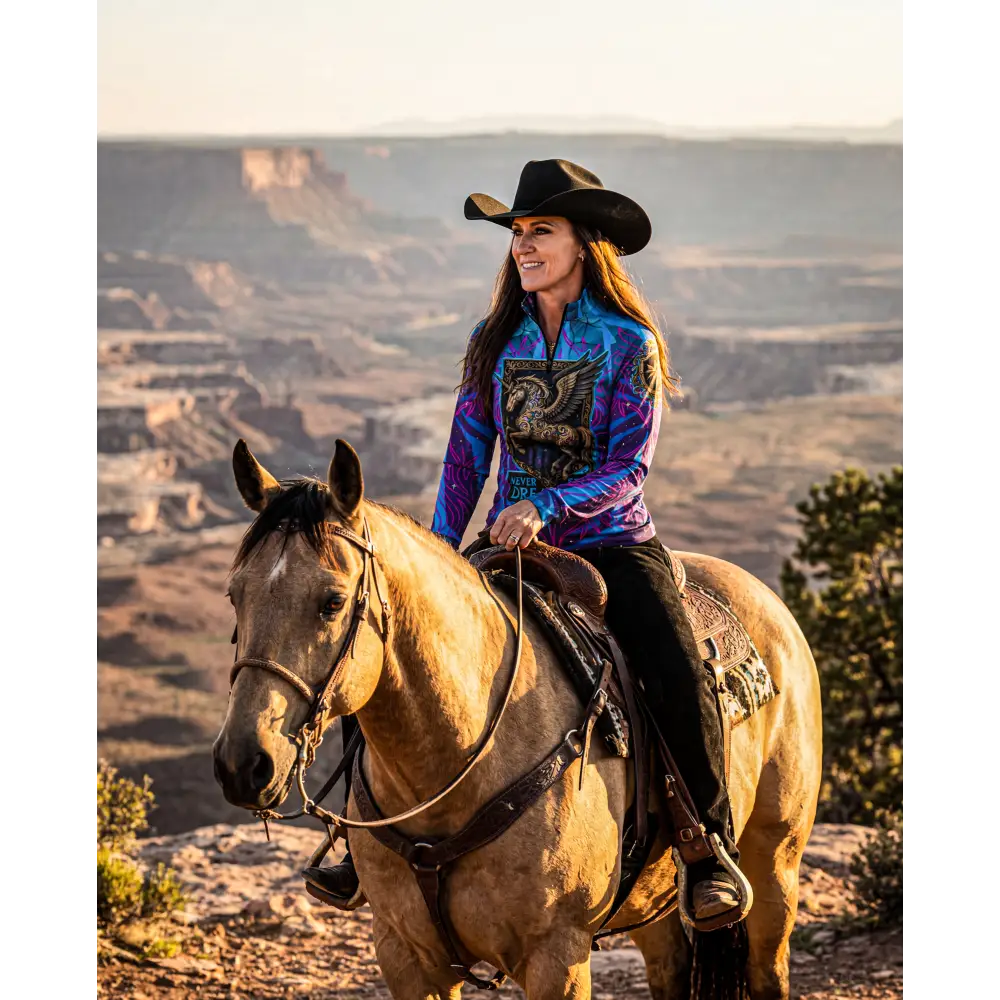 A woman wearing a vibrant blue and purple never stop dreaming performance equestrian sun shirt with upf50+ paired with black pants and a cowboy hat