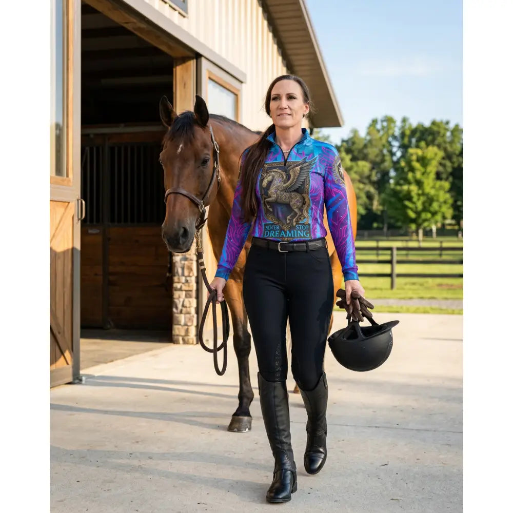The woman wears a vibrant long-sleeved performance equestrian sun shirt with a winged horse and stop dreaming print