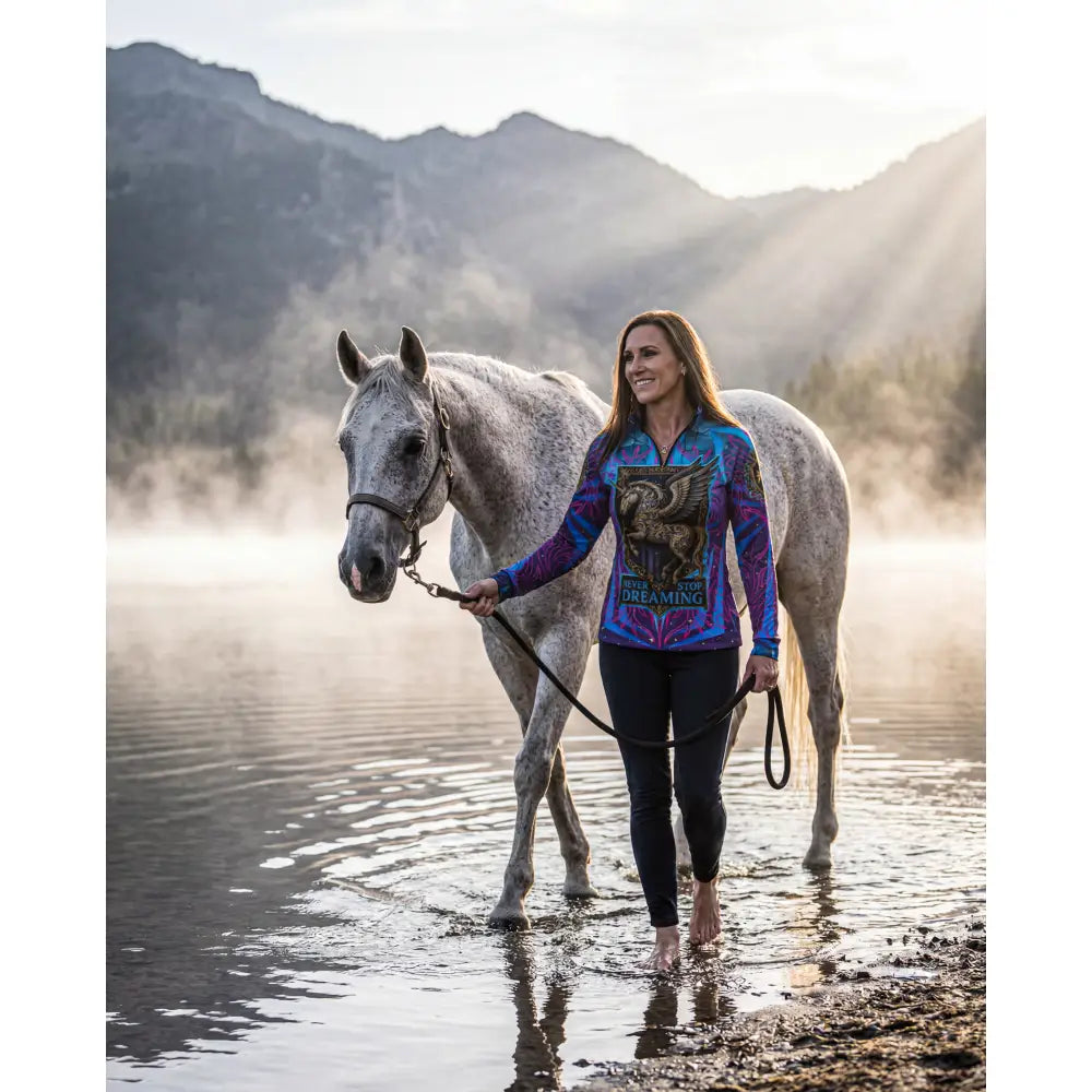 A woman walks barefoot beside a gray horse wearing a vibrant winged horse print performance equestrian sun shirt