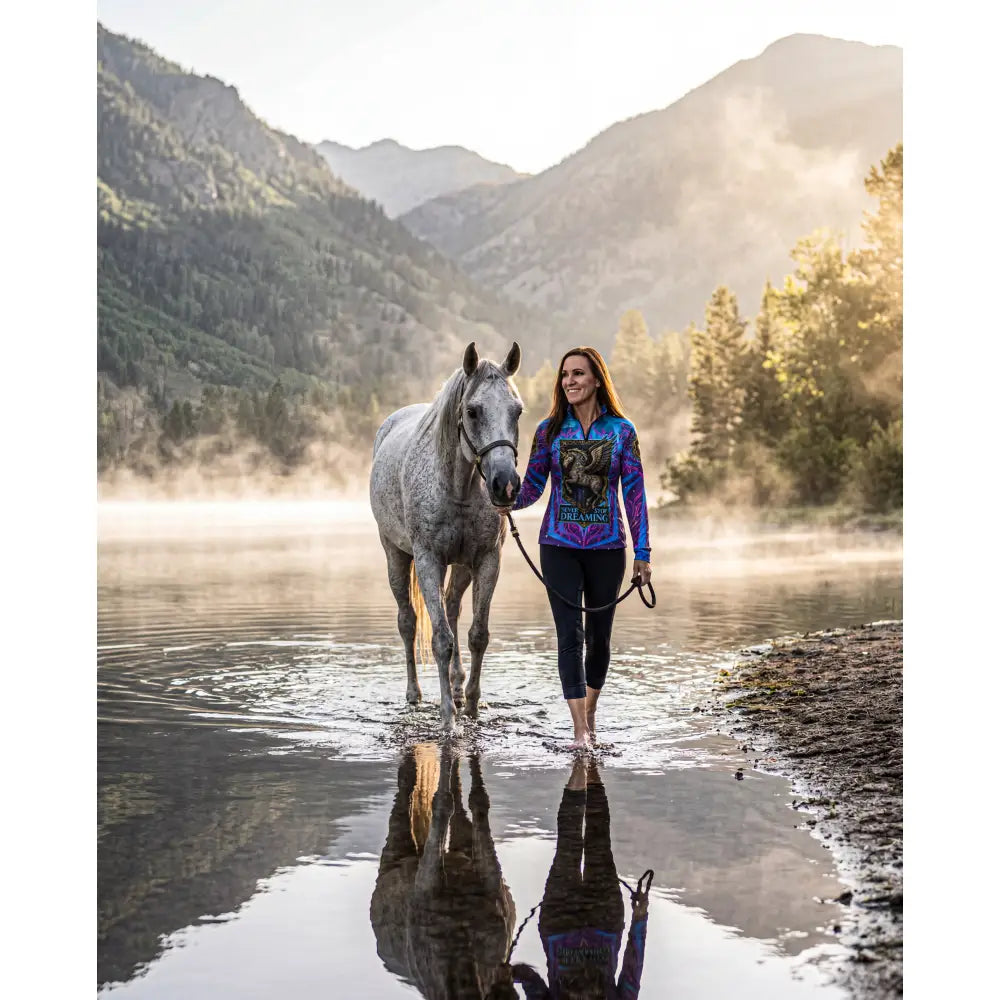 A woman walks barefoot beside a gray horse wearing a vibrant dragon print equestrian sun shirt stop dreaming design