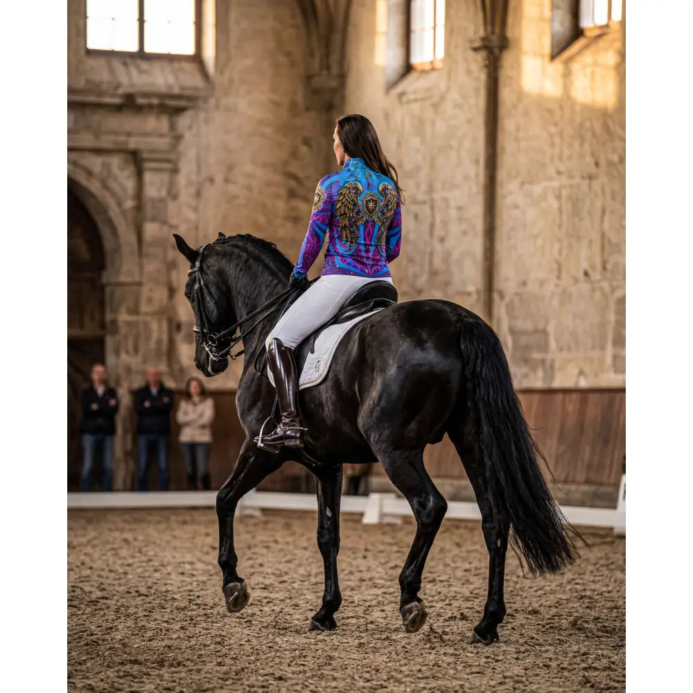 A rider in a vibrant purple and blue performance equestrian sun shirt with golden winged designs atop a sleek black horse