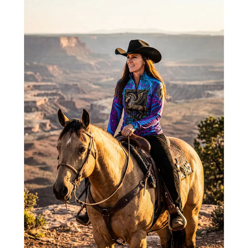 A woman wearing a vibrant blue and purple never stop dreaming equestrian sun shirt with embroidered dragon design and black cowboy hat