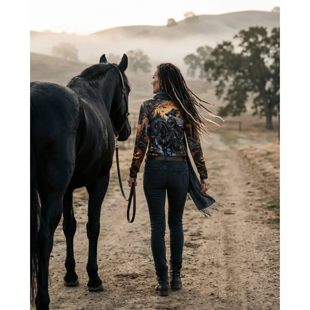 A woman with long dreadlocks walks beside a black horse wearing púca’s eve dark horse witch equestrian sun shirt by karen reinertson