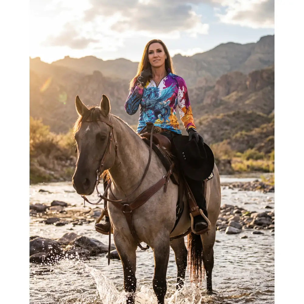 A woman rides a gray horse through shallow water wearing the vibrant riptide radiance upf50+ equestrian sun shirt