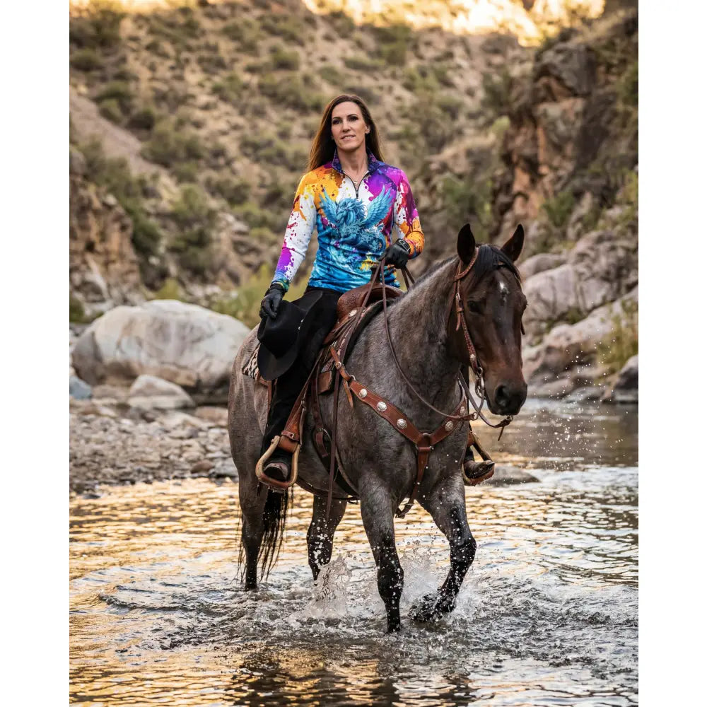 A woman rides a dark gray horse wearing the riptide radiance wearable art upf50+ equestrian sun shirt from the ripley signature collection