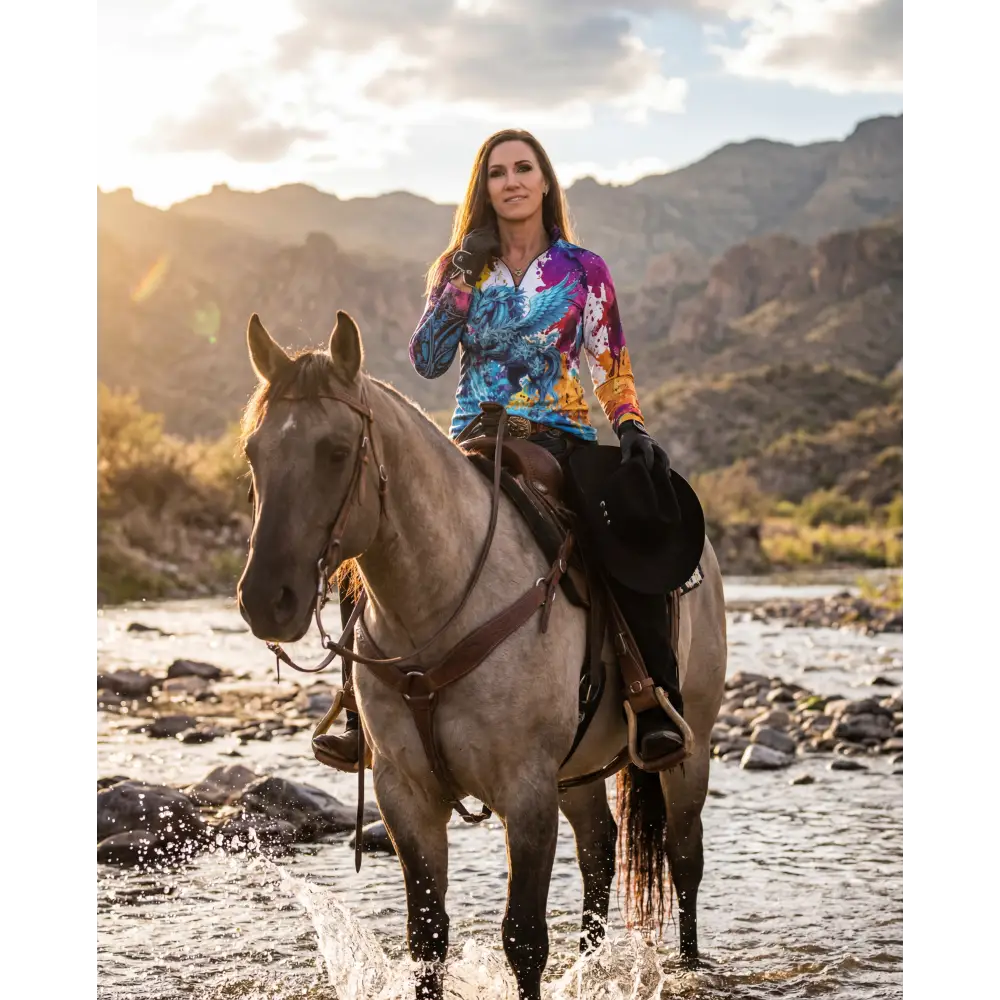 A woman rides a gray horse through shallow water wearing a vibrant tie-dye riptide radiance sun shirt upf50+
