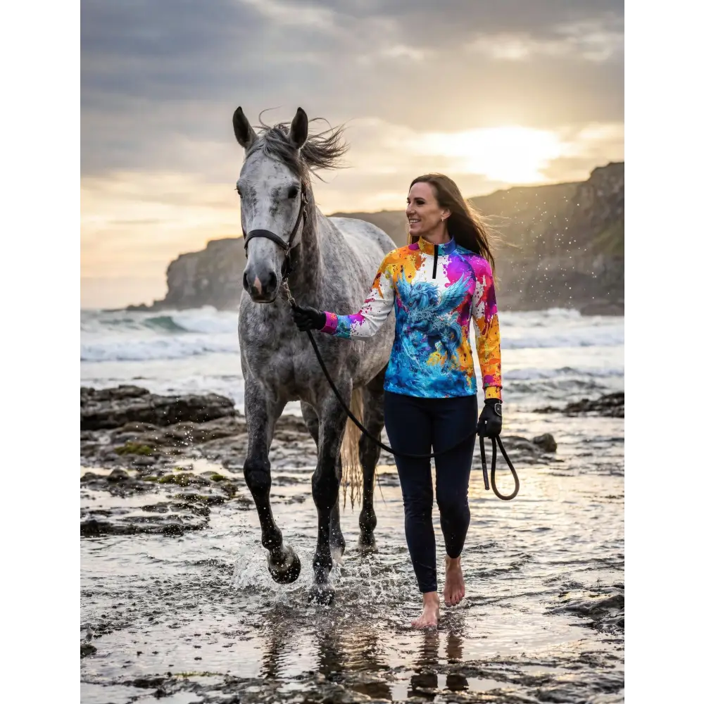 A woman walks barefoot along the shore with her grey horse wearing a vibrant tie-dye riptide radiance sun shirt upf50+