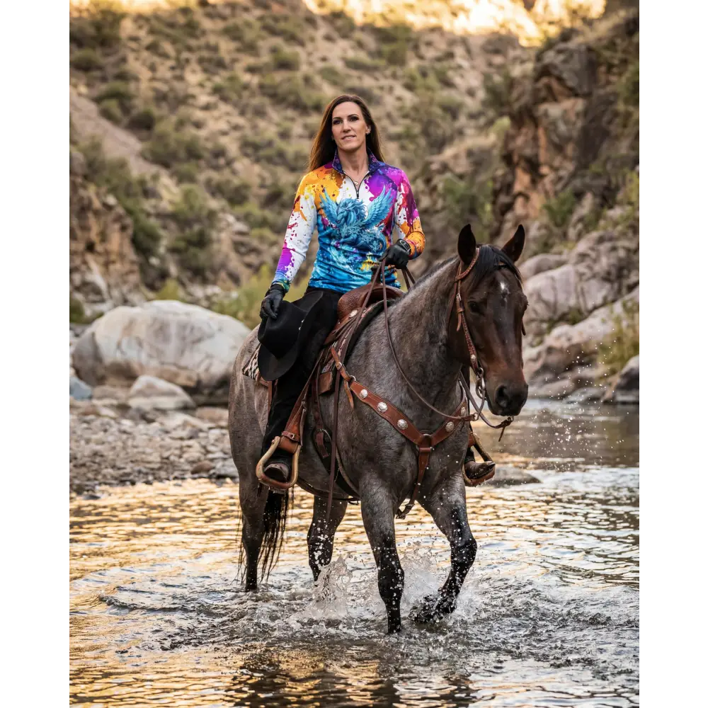 A woman rides a dark gray horse wearing a vibrant tie-dye riptide radiance performance equestrian sun shirt upf50+