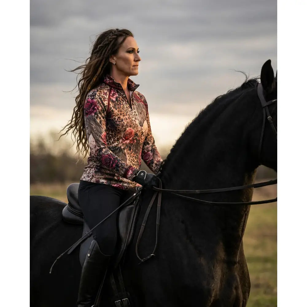 A woman rides a black horse wearing karen reinertsons rosae equiferus leopard rose equestrian sun shirt with upf50+