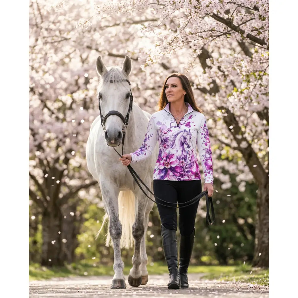A woman wearing a purple floral zip-front sakura wildheart equestrian sun shirt walks beside a gray horse under cherry trees