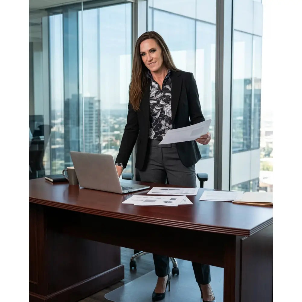 Woman in black blazer and gray trousers standing at wooden desk wearing shadow bloom performance dark floral print upf50+ equestrian sun shirt with cooling fabric