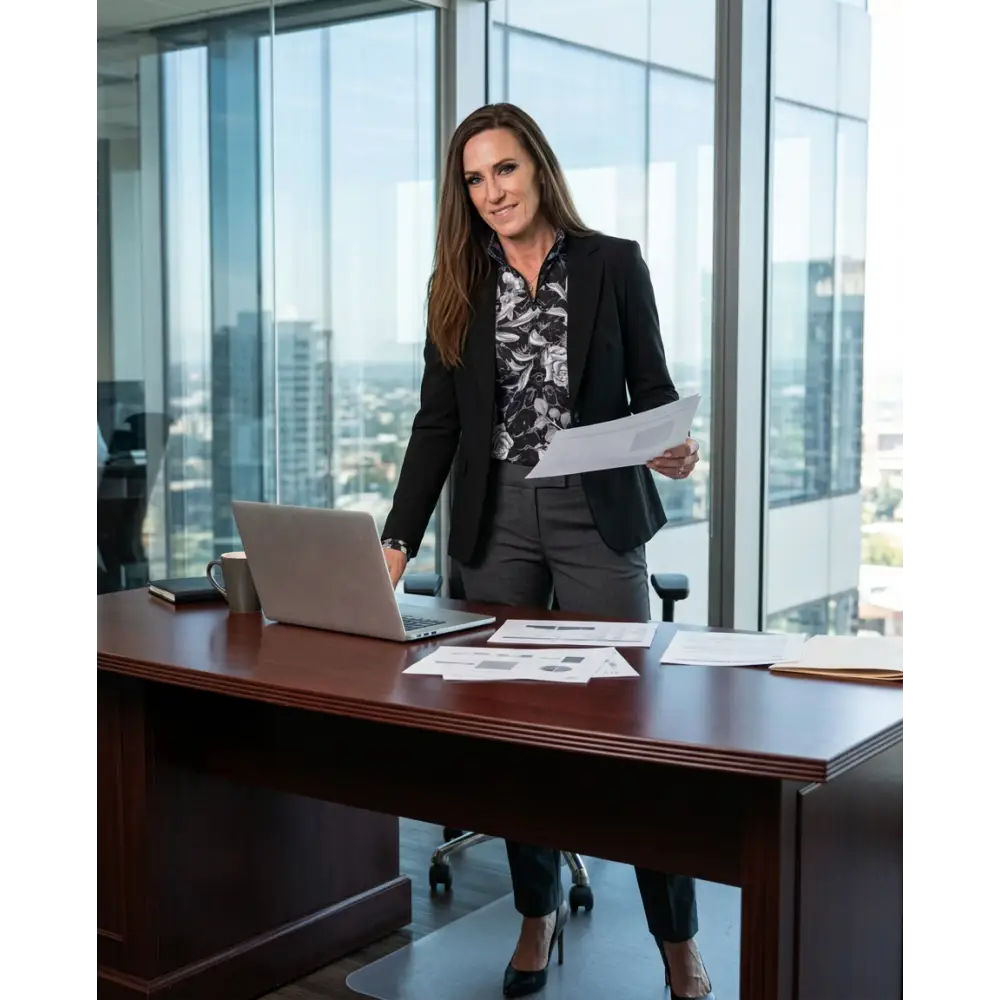 Woman in black blazer and gray trousers standing at wooden desk with papers and laptop showing shadow bloom sun shirt upf50+
