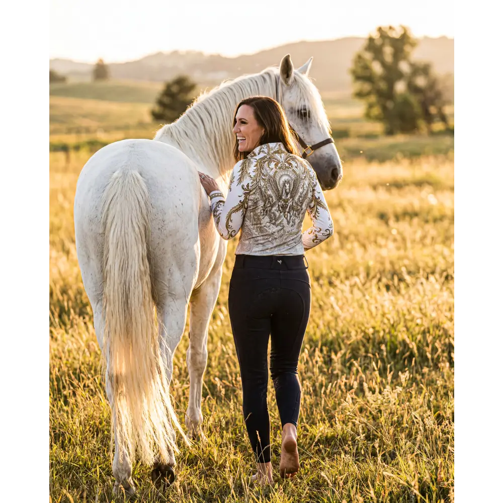 A woman wears a white and gold embroidered solis aeternum performance equestrian sun shirt upf50+ beside a white horse