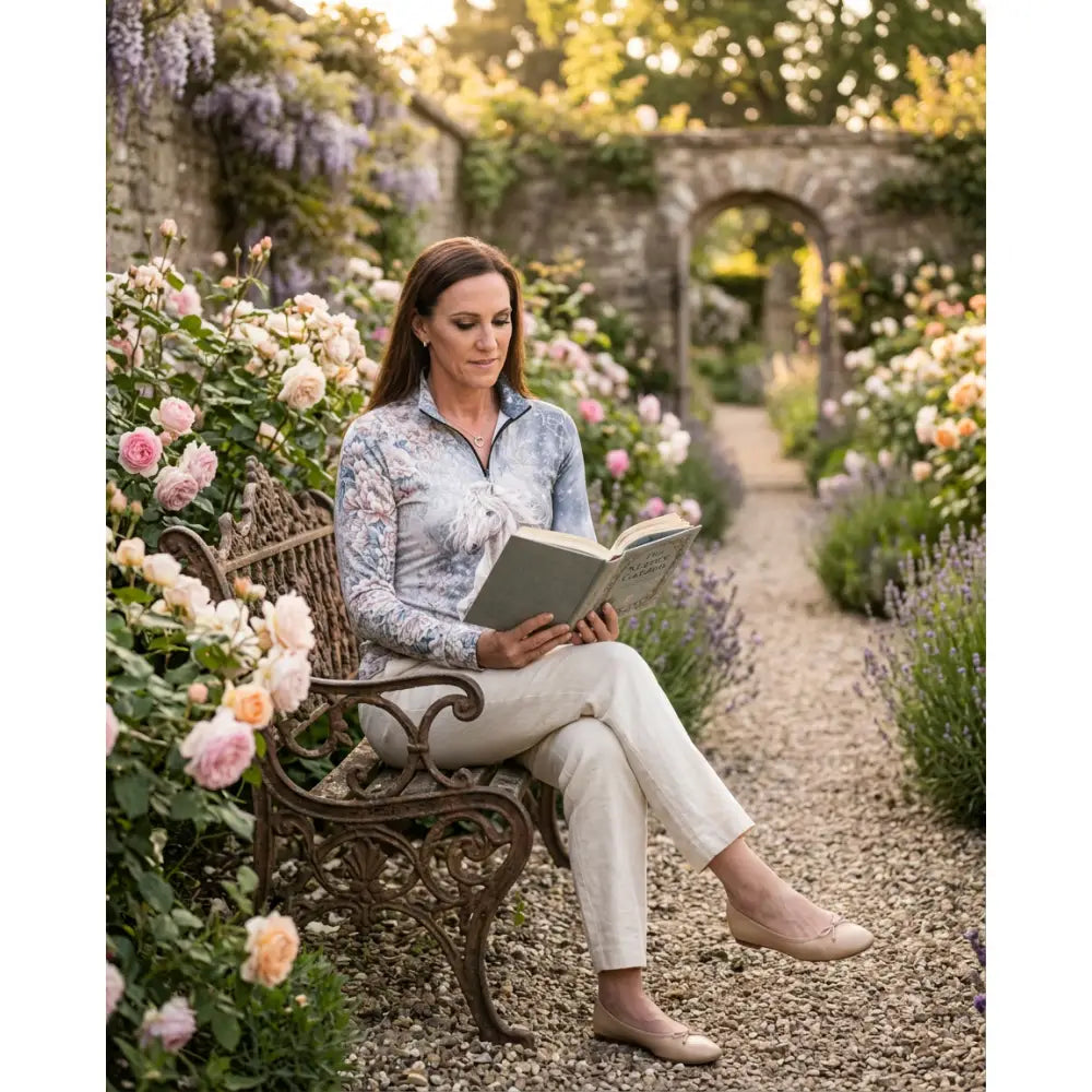 Woman wearing unicornis caelestis equestrian sun shirt in light blue floral print seated on wrought iron bench in garden