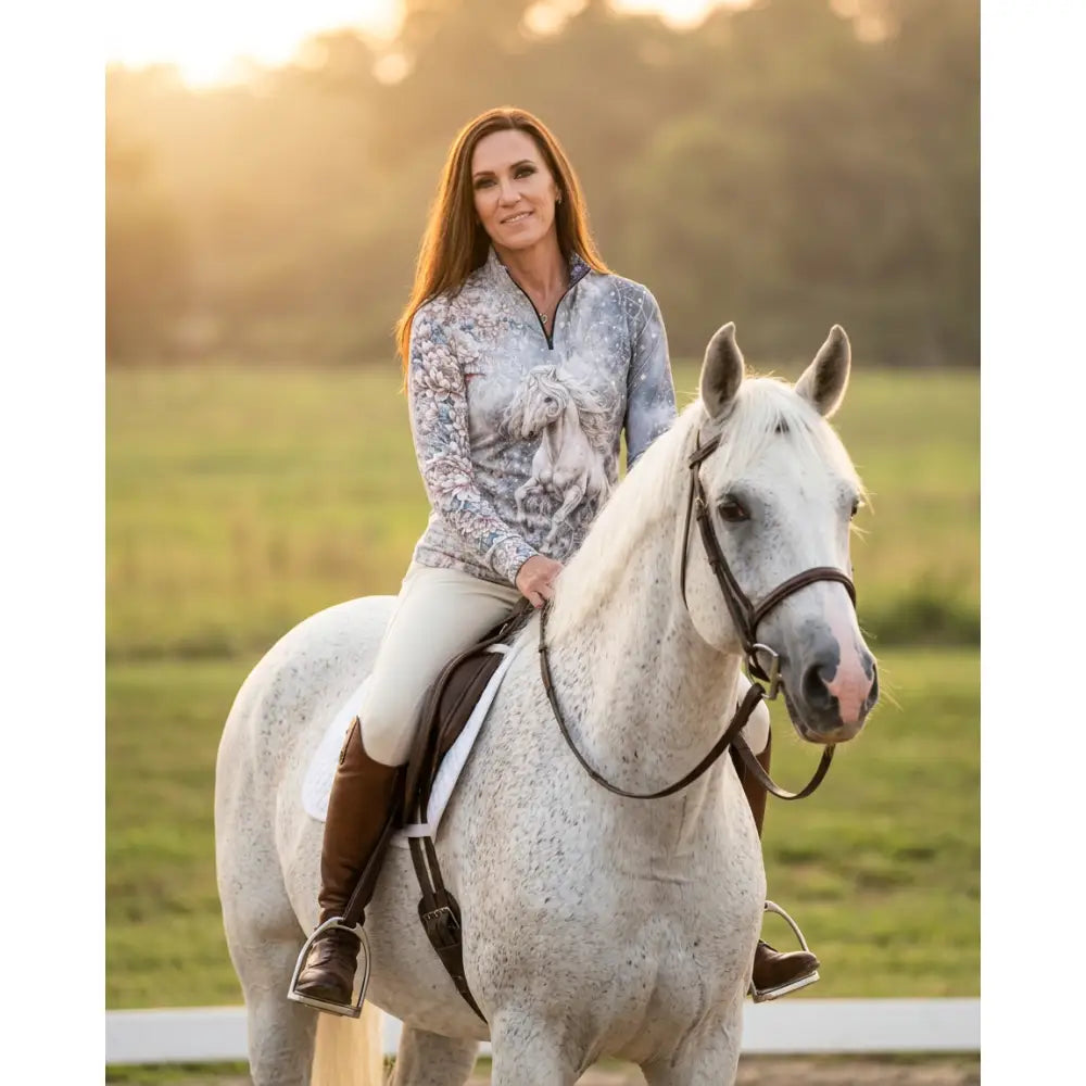 A woman wearing a light blue and gray unicornis caelestis equestrian sun shirt with a white horse graphic by karen reinertson