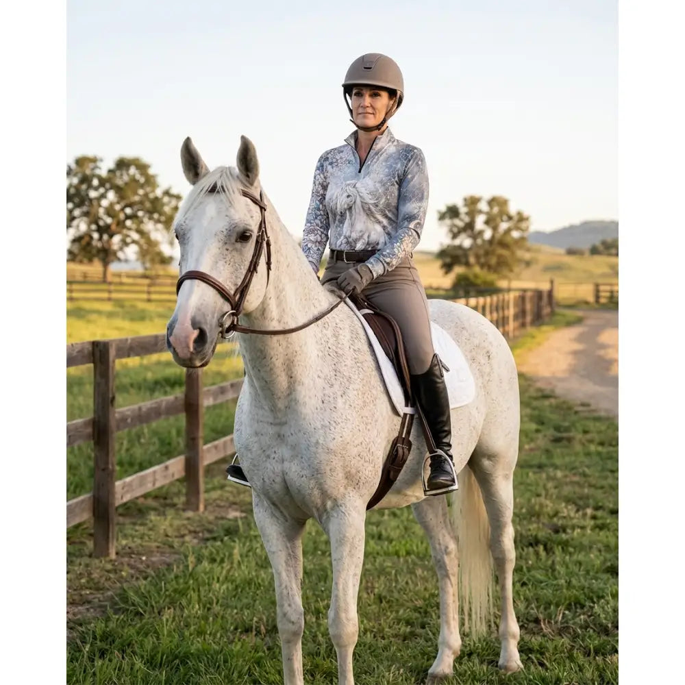 A woman wearing karen reinertson unicornis caelestis equestrian sun shirt rides a white speckled horse in a helmet