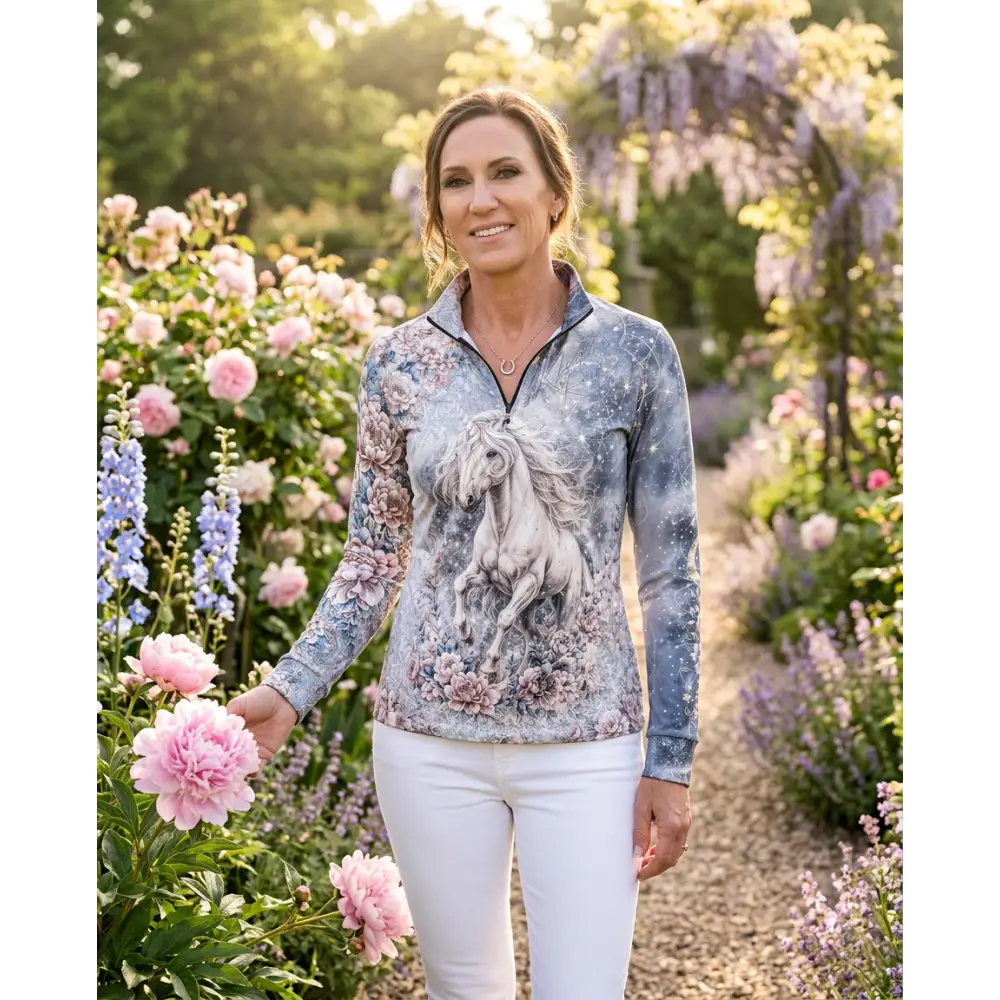 A woman wears a unicornis caelestis performance equestrian sun shirt upf50 plus with white horse print on blue gray fabric