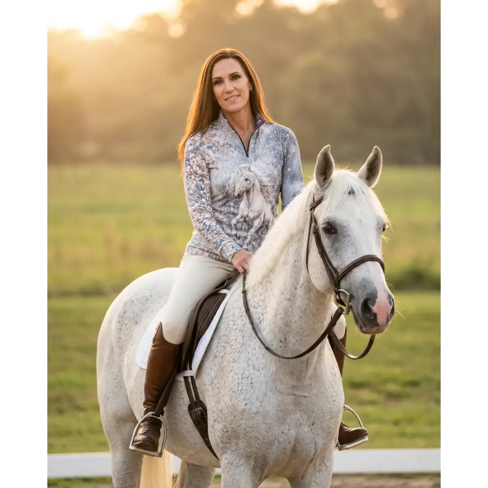 A woman wearing a light blue gray unicornis caelestis performance equestrian sun shirt with a white horse graphic and UPF50+