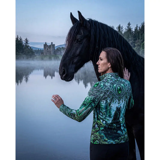 A woman wears a whispering waters riding shirt by karen reinertson with a green black forest design and emerald accents