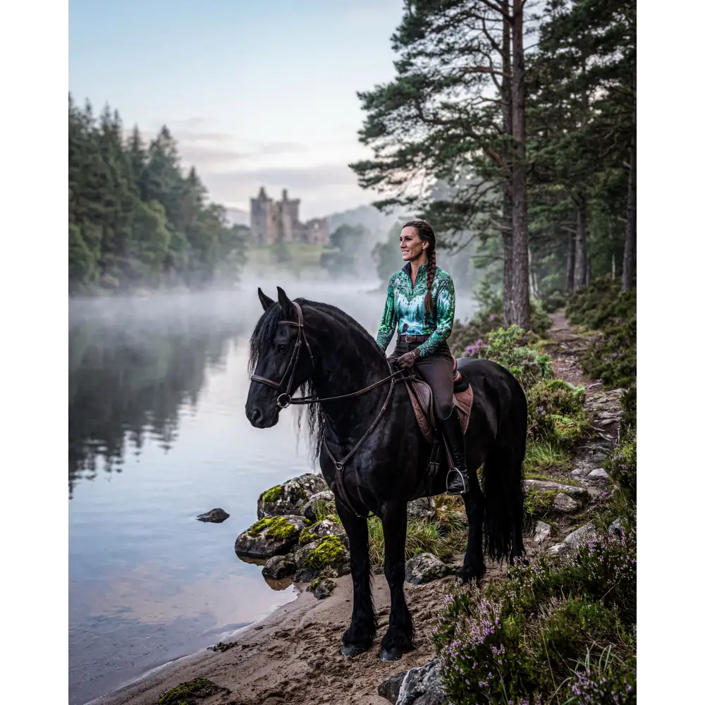 A woman in a teal and green patterned whispering waters performance equestrian sun shirt with upf50 plus rides a black horse near misty water