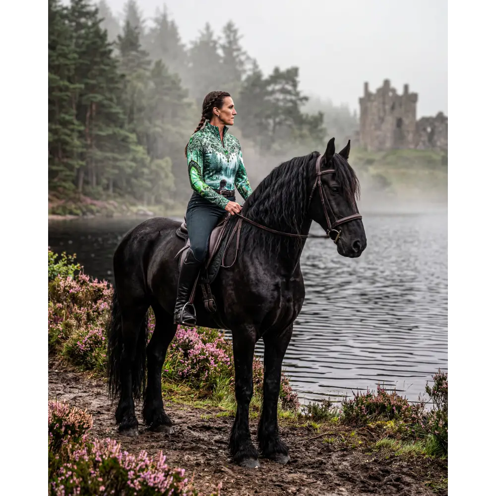 A rider wears a green and black patterned whispering waters performance equestrian sun shirt upf50 plus on a black horse