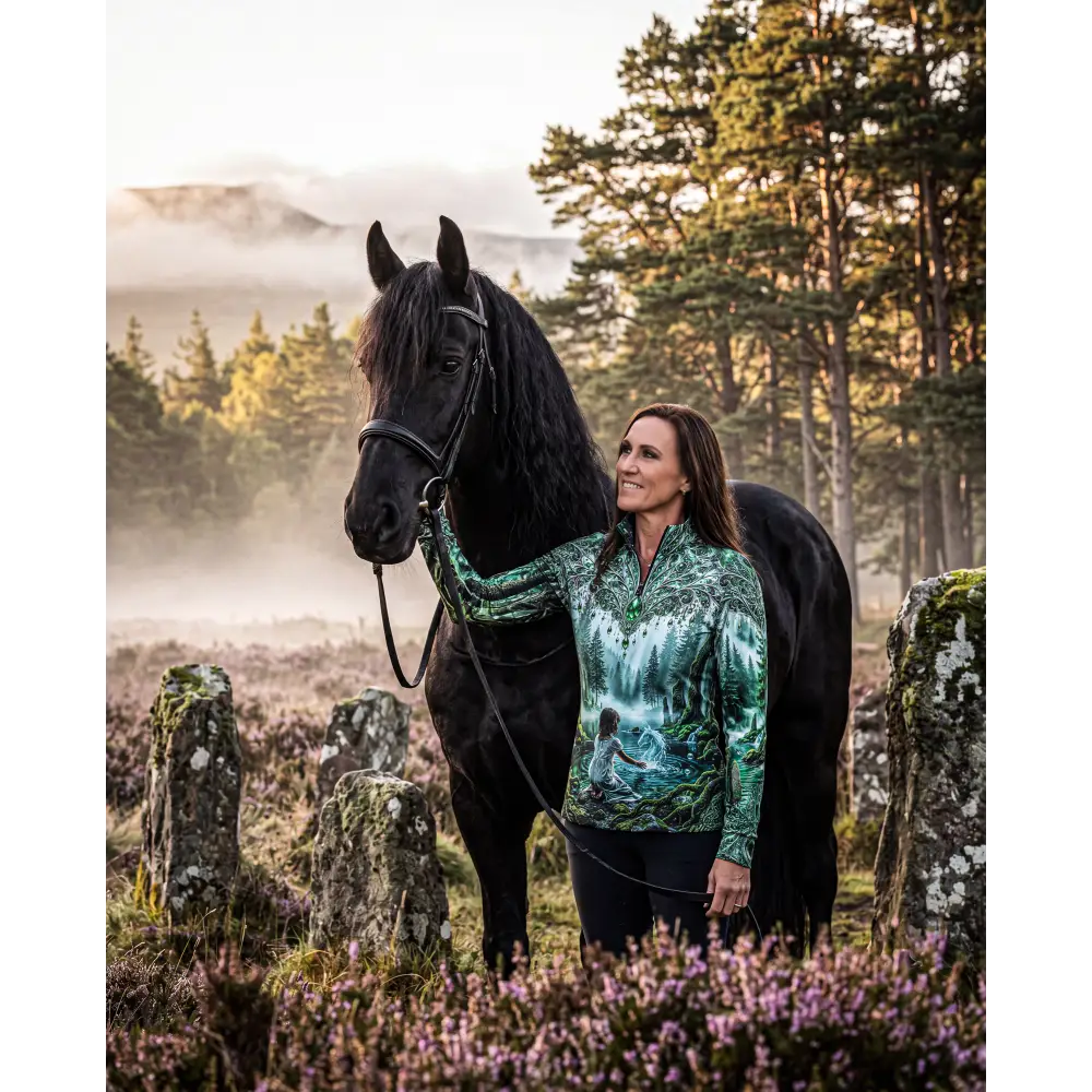 A woman wearing a forest-themed whispering waters performance equestrian sun shirt upf50 plus stands by a black horse