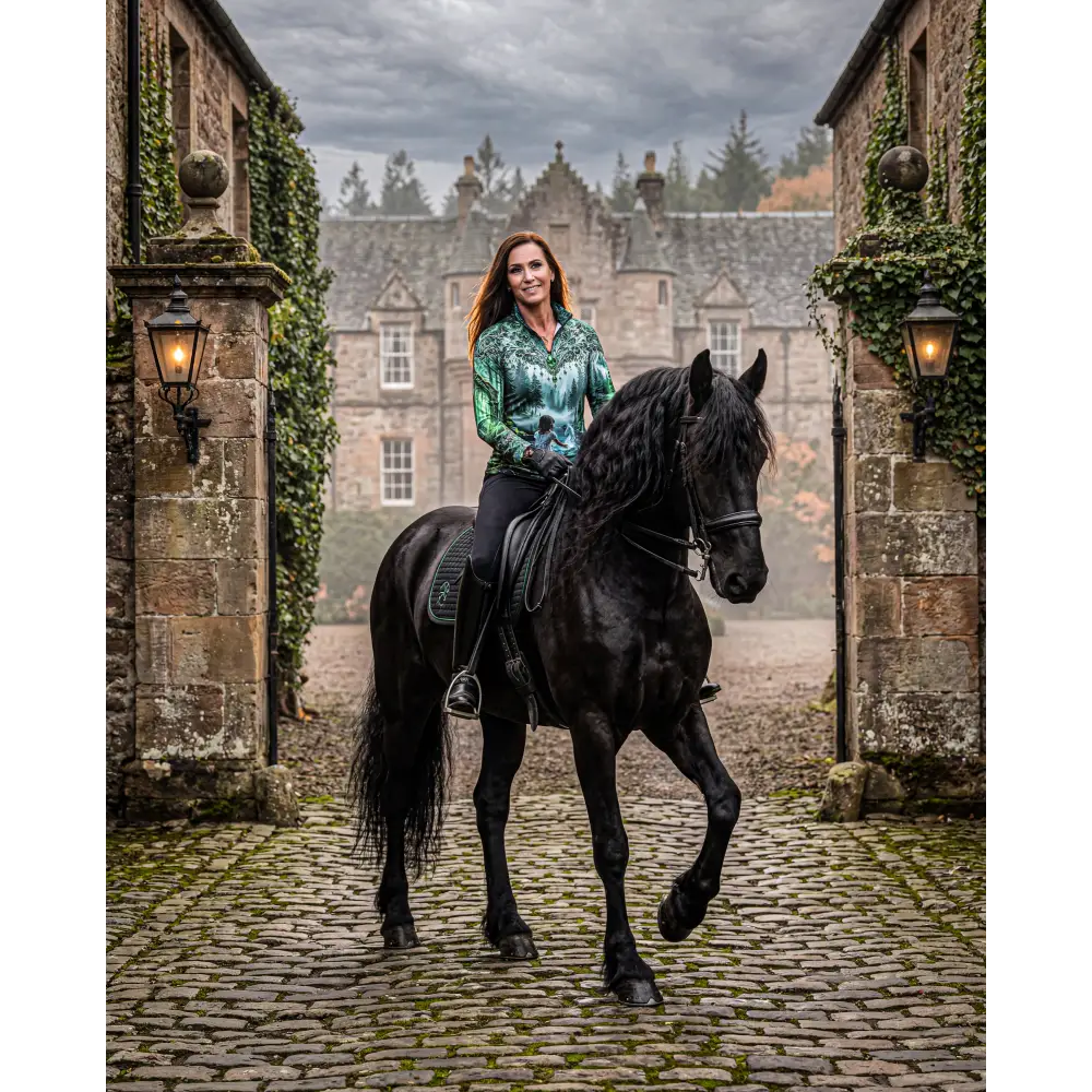 A woman in a green patterned whispering waters performance equestrian sun shirt upf50+ rides a black friesian horse